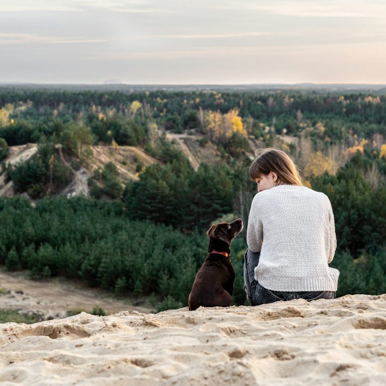 Frau mit Hund Eine Person sitzt mit einem Hund auf einer Sanddüne, sie schauen sich an.
