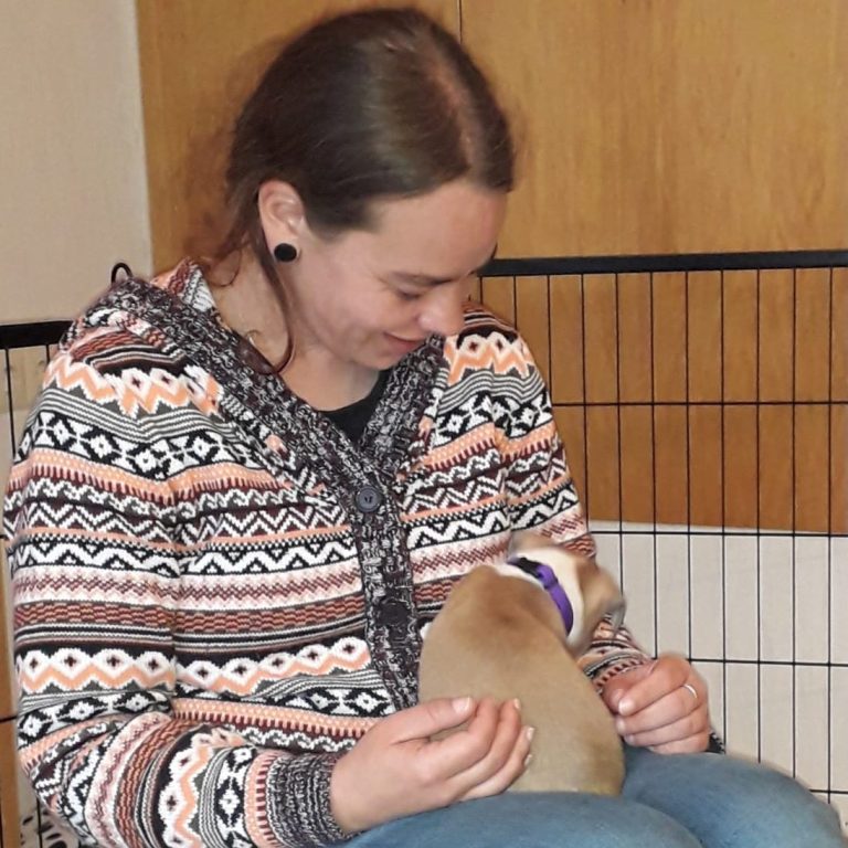 K. Kassner Eine Frau mit langen, dunkelblonden Haaren, die eine gemusterte Strickjacke anhat, hält einen Welpen im Schoß, und schaut diesen an